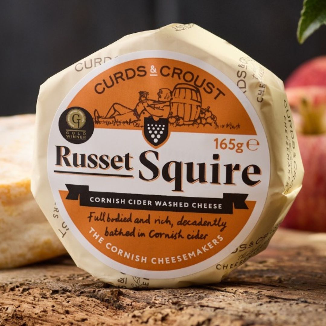 Russet Squire Cornish cider-washed brie by Curds & Croust, whole round with distinctive russet rind, on a wooden board — available from The Cornish Food Box Company.