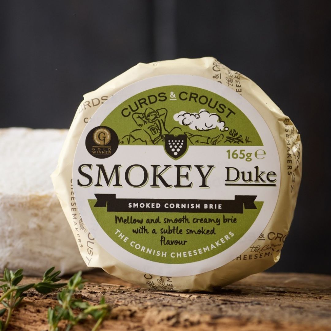 Smokey Duke oak-smoked Cornish brie by Curds & Croust, whole round with velvety rind, on a wooden board — available from The Cornish Food Box Company.